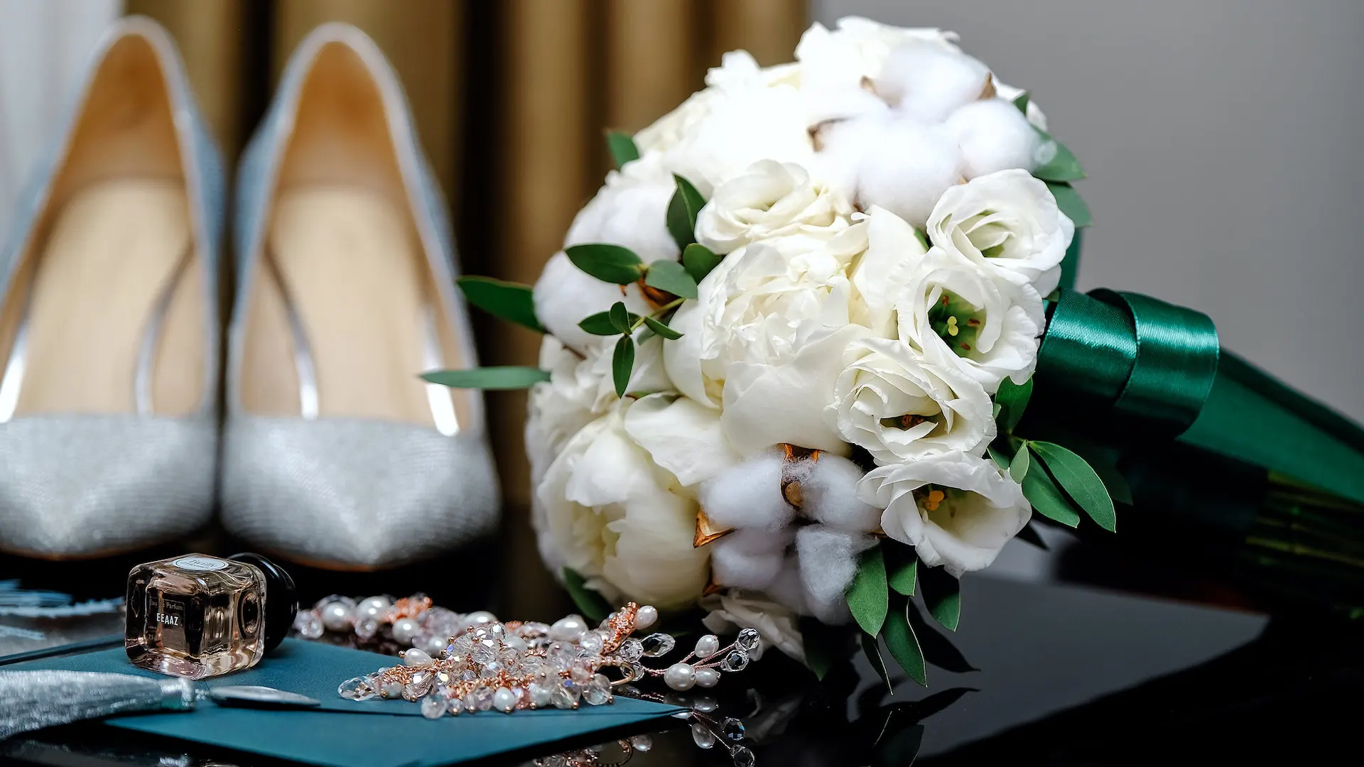 Close up of silver women's pump shoes, bouquet of white flowers, earrings, and perfume sitting on a table ready for a special event