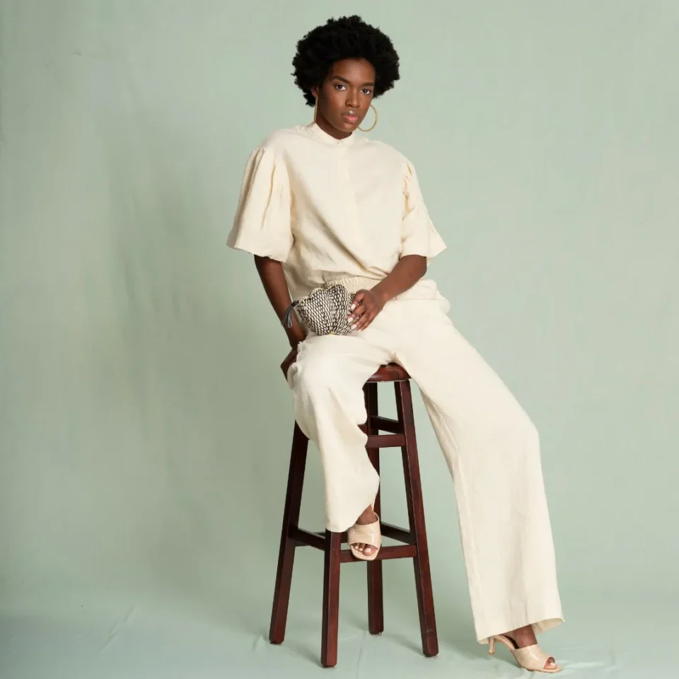 young black woman with a stylish afro wearing cream colored fashion jumpsuit sitting on stool holding her bag